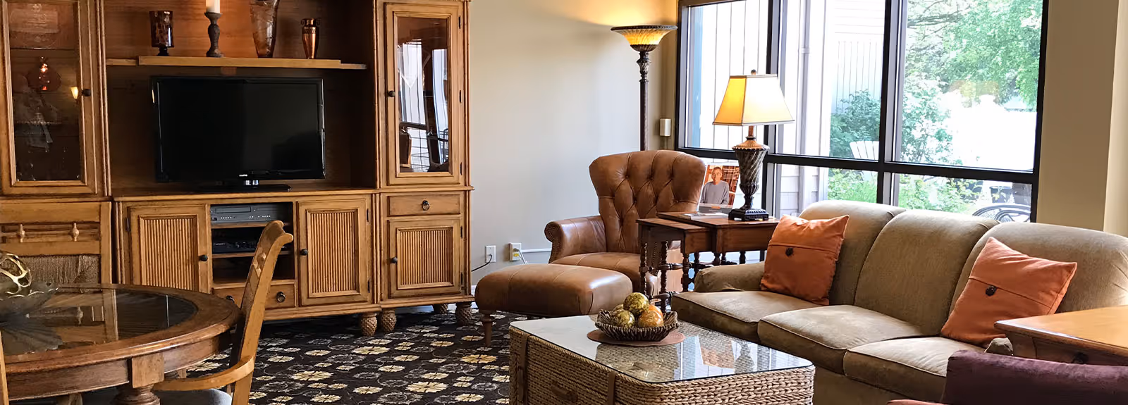 A cozy living room with a beige sofa adorned with orange pillows, a brown leather armchair with matching ottoman, a wooden coffee table with decorative items, a wooden entertainment center with a TV, and large windows letting in natural light.