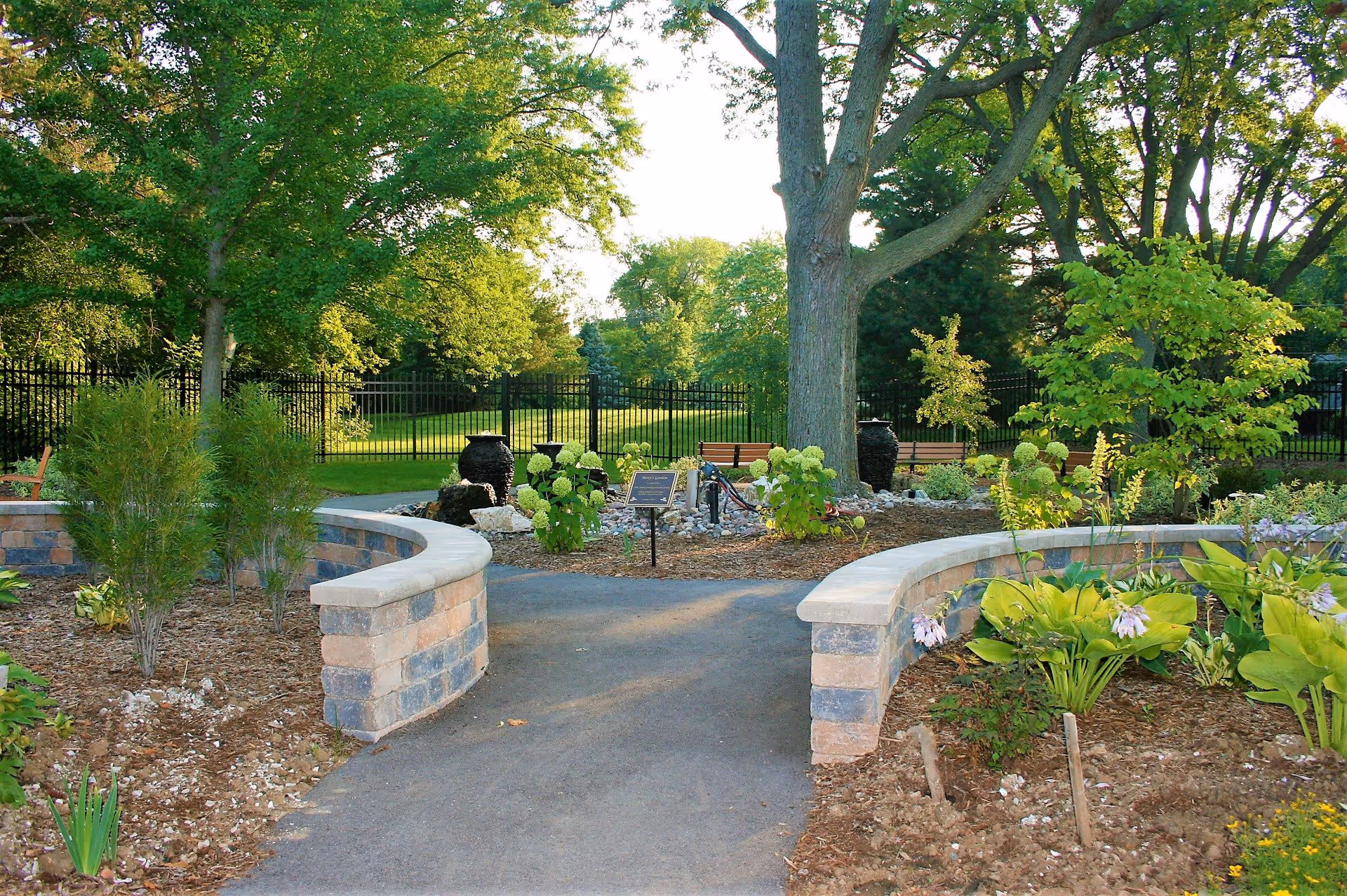 A peaceful outdoor garden area with a paved pathway flanked by low stone walls and various green plants and flowers. Large trees provide shade, and there are benches and decorative urns in the background near a black metal fence.