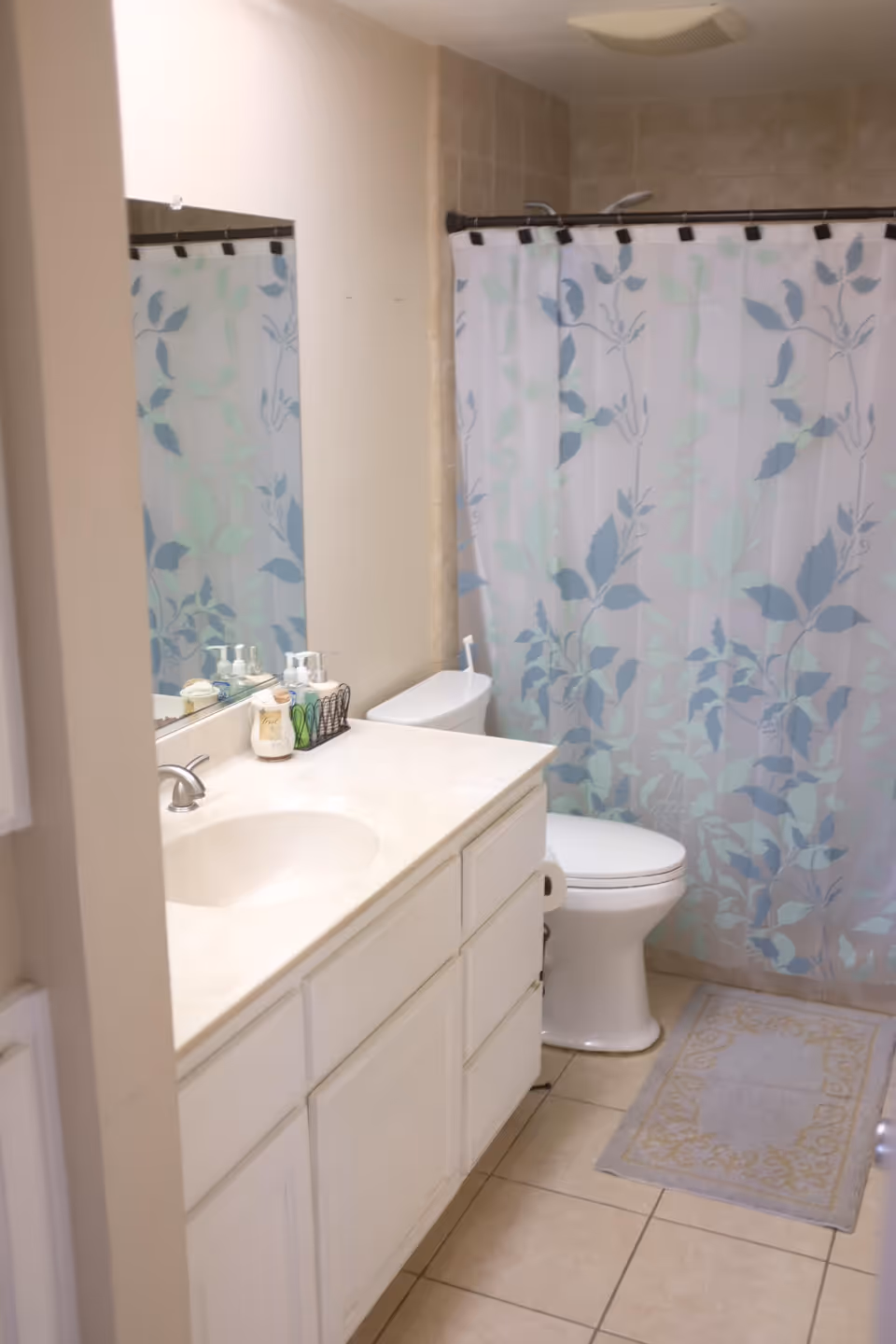 A clean bathroom with a white vanity and sink, a large mirror above the sink, a toilet next to the vanity, and a shower with a floral-patterned shower curtain. The floor is tiled and there is a small decorative rug in front of the shower.