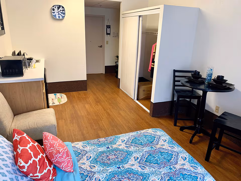Interior view of a supportive living facility room showing a bed with blue and white patterned bedding and red pillows, a small beige couch, a kitchenette area with a microwave, a round black dining table set for two, and an open closet with sliding doors. The floor is wood laminate and the walls are white.
