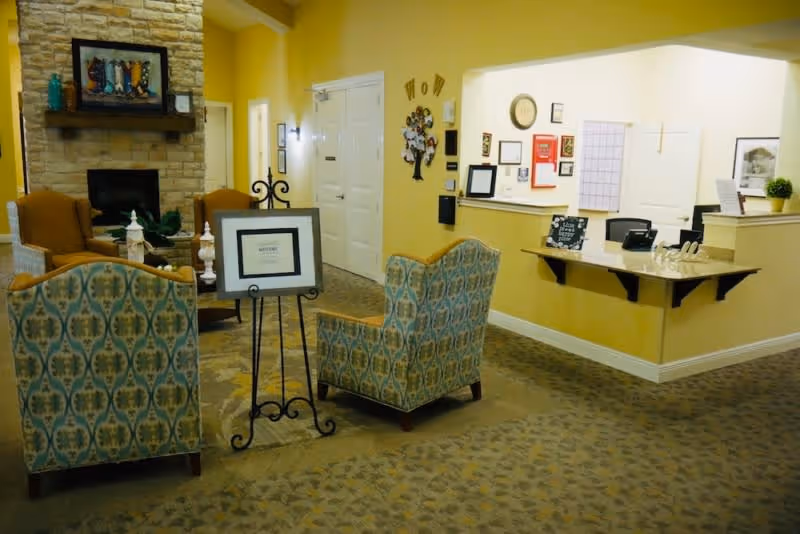A cozy senior living facility common area with patterned armchairs, a stone fireplace, and a reception desk. The walls are painted yellow, and there are various framed pictures and decorations on the walls. The carpet has a subtle pattern, and the space is warmly lit.