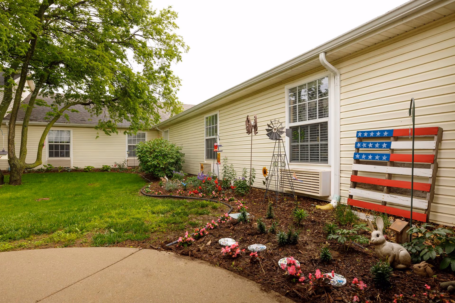 A garden area outside a beige building with green grass, a tree, and a flower bed containing various plants, garden decorations including a small windmill, a wooden American flag, and a rabbit statue.