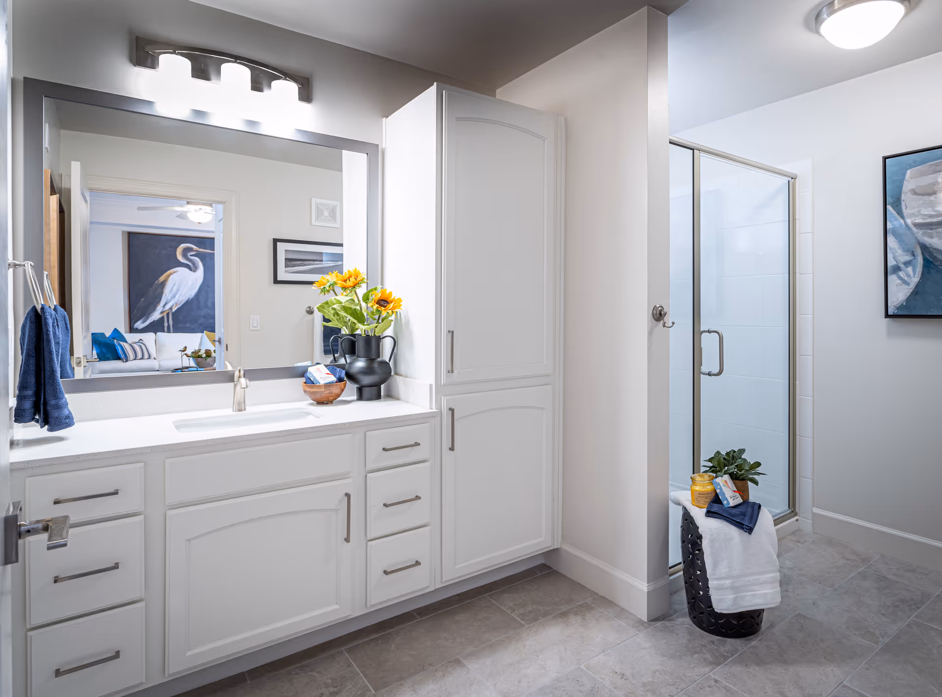 Bright and clean bathroom with white cabinetry, a large mirror above the sink, a black vase with sunflowers on the countertop, and a glass-enclosed shower. A small black stool holds a white towel, a plant, and a yellow jar. The reflection in the mirror shows a room with a large bird painting and a white couch with blue and yellow pillows.
