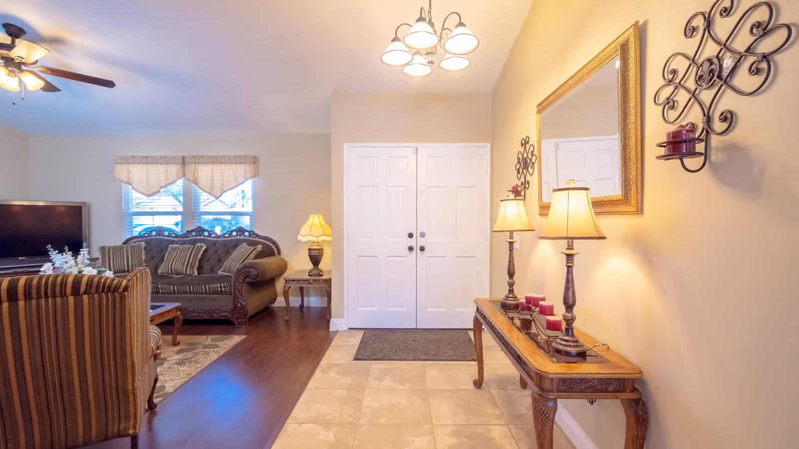 A cozy living room area with a striped armchair and a dark green sofa with multiple cushions. There is a wooden coffee table and a TV on a stand near two windows with beige valances. The entryway features a double white door, a wooden console table with two lamps, red candles, and a large framed mirror above it. The walls are light beige, and the floor transitions from tiled near the door to wood in the living area.