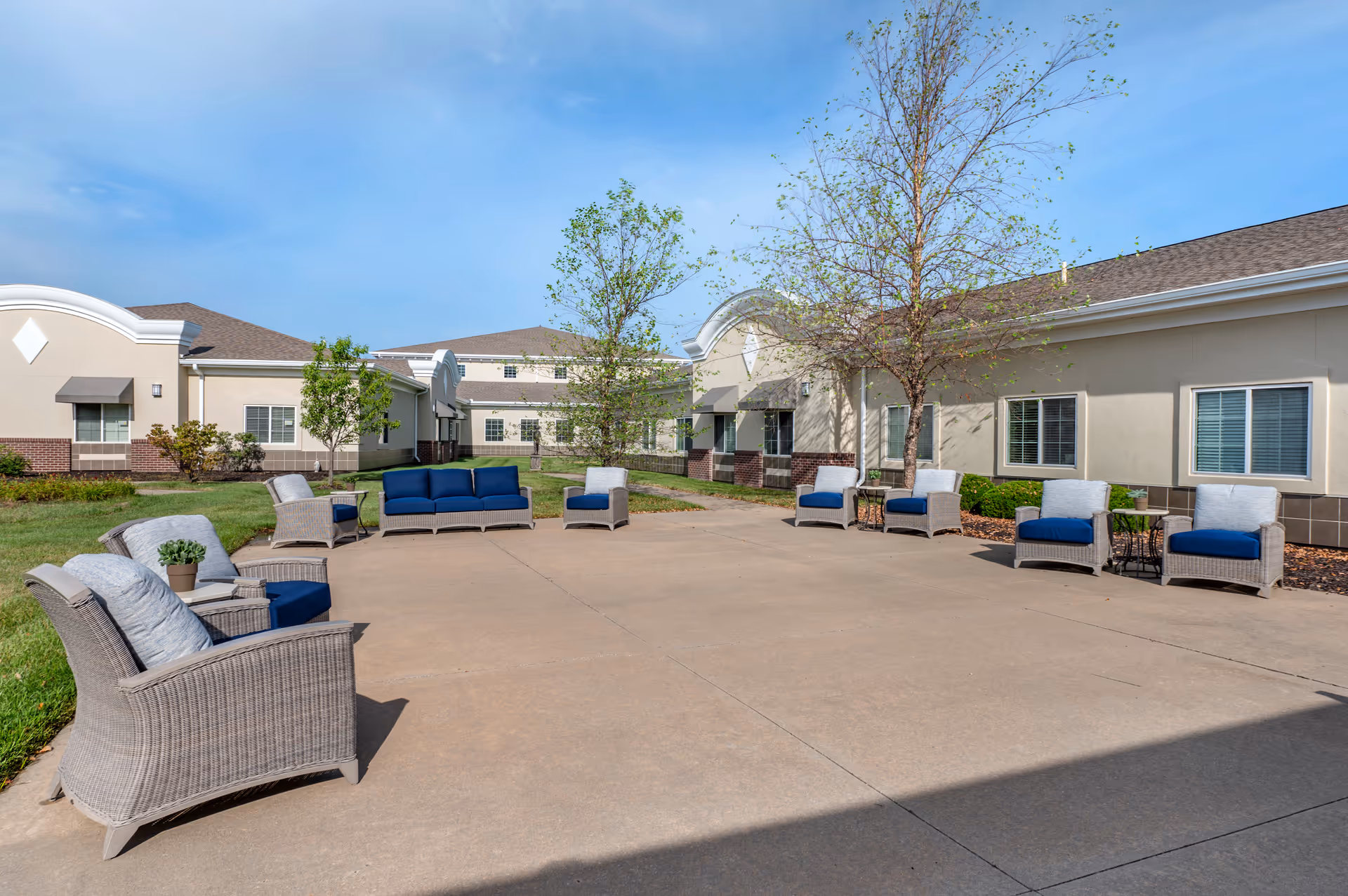 Outdoor courtyard with wicker seating and blue cushions in front of a single-story senior living facility building.
