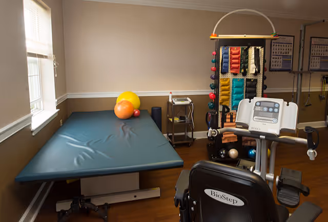 A small physical therapy or exercise room with a padded treatment table holding exercise balls, a rack with various weighted bags and balls, and a BioStep exercise machine. The room has beige walls with white trim and a window letting in natural light.