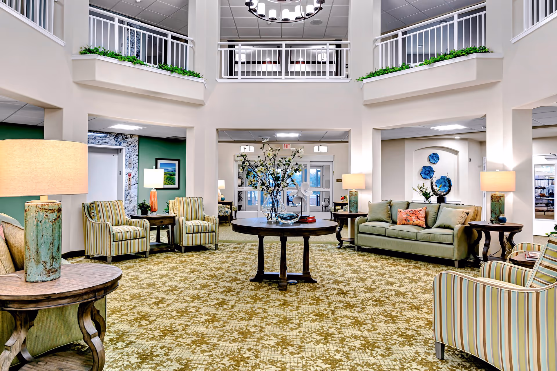 A spacious and well-lit living room area in a senior living facility with a high ceiling and balcony railing above. The room features a patterned carpet, a round wooden table with a floral arrangement in the center, several upholstered chairs with striped fabric, a green sofa with decorative pillows, and multiple side tables with lamps. The walls are light-colored with some green accent walls and decorative blue wall art.