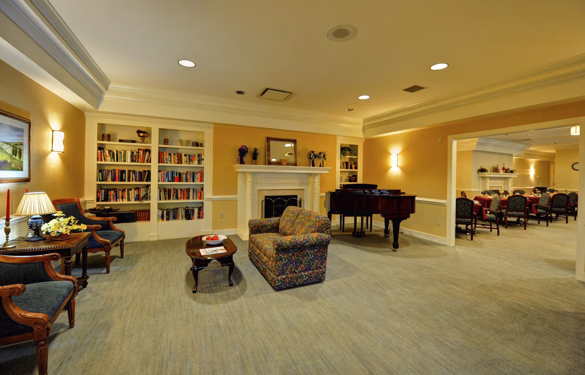 A cozy living room area in a senior living facility featuring a floral patterned armchair, a wooden coffee table with a bowl of apples and magazines, two upholstered wooden chairs, built-in bookshelves filled with books, a white fireplace with decorative items on the mantel, and a grand piano. The room has warm yellow walls, soft lighting, and carpeted flooring. In the background, there is a dining area with tables and chairs set up.