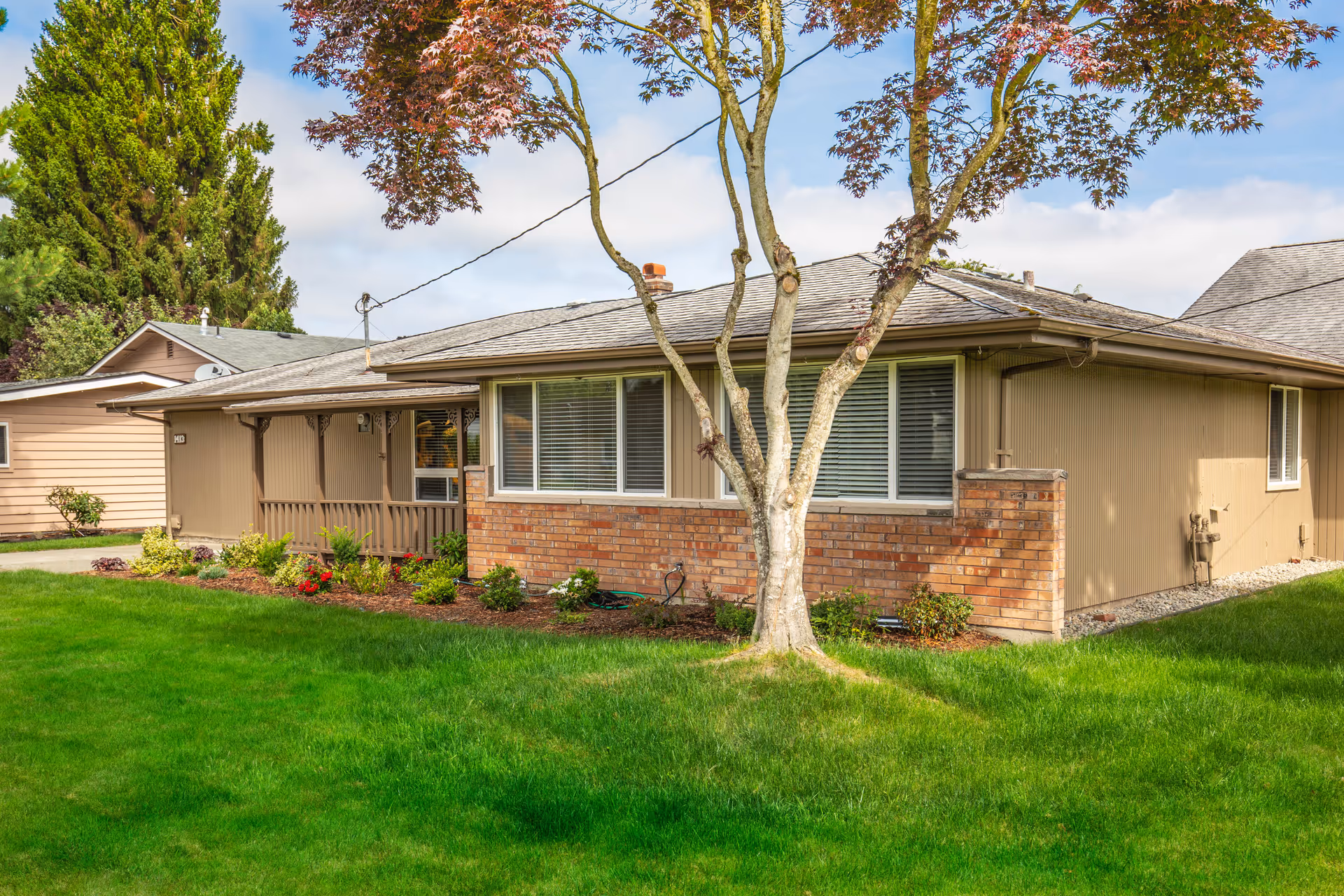 Exterior view of a single-story residential building with beige siding and a brick lower facade. There is a small porch area with decorative wooden supports, a well-maintained green lawn, a tree with reddish leaves in the foreground, and some shrubs and flowers planted along the building's foundation. The sky is partly cloudy.