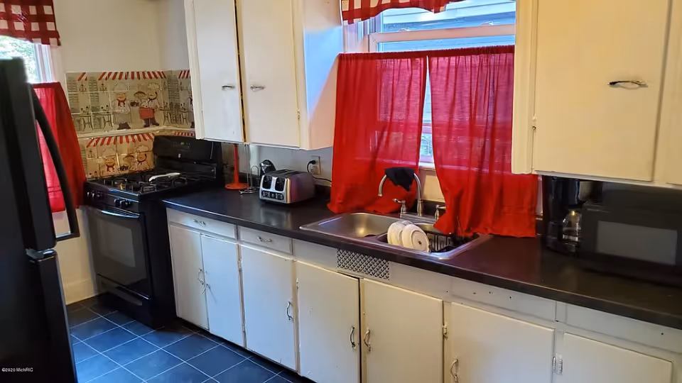 A kitchen with white cabinets, black countertops, and black appliances including a stove, toaster, microwave, and coffee maker. There is a window above the sink with red curtains, and a dish rack with dishes in the sink. The floor is tiled in black, and there is a decorative backsplash with a chef motif behind the stove.