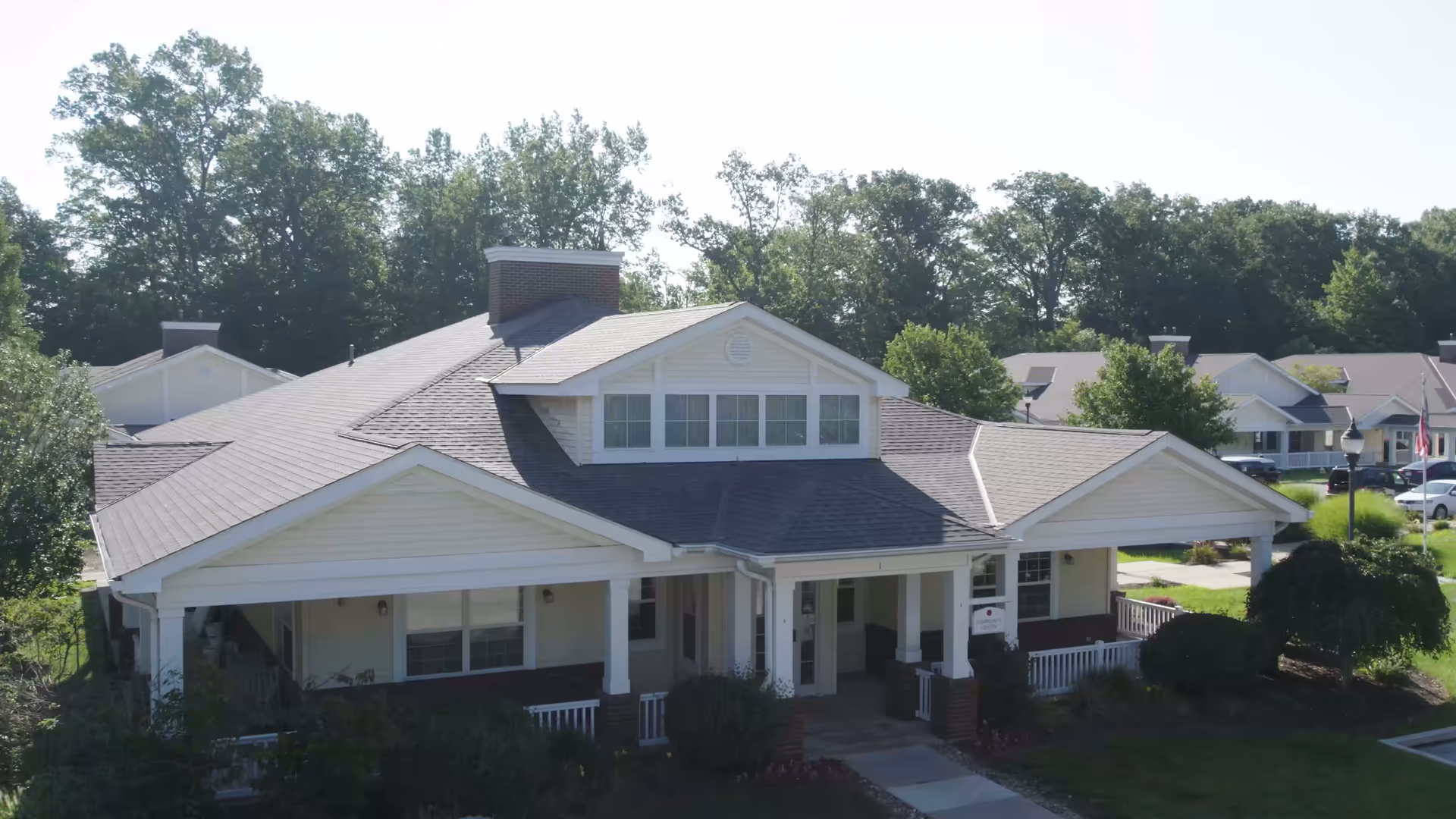 Exterior view of a single-story senior living facility building with a covered entrance, white siding, and a gray shingled roof surrounded by greenery and trees.