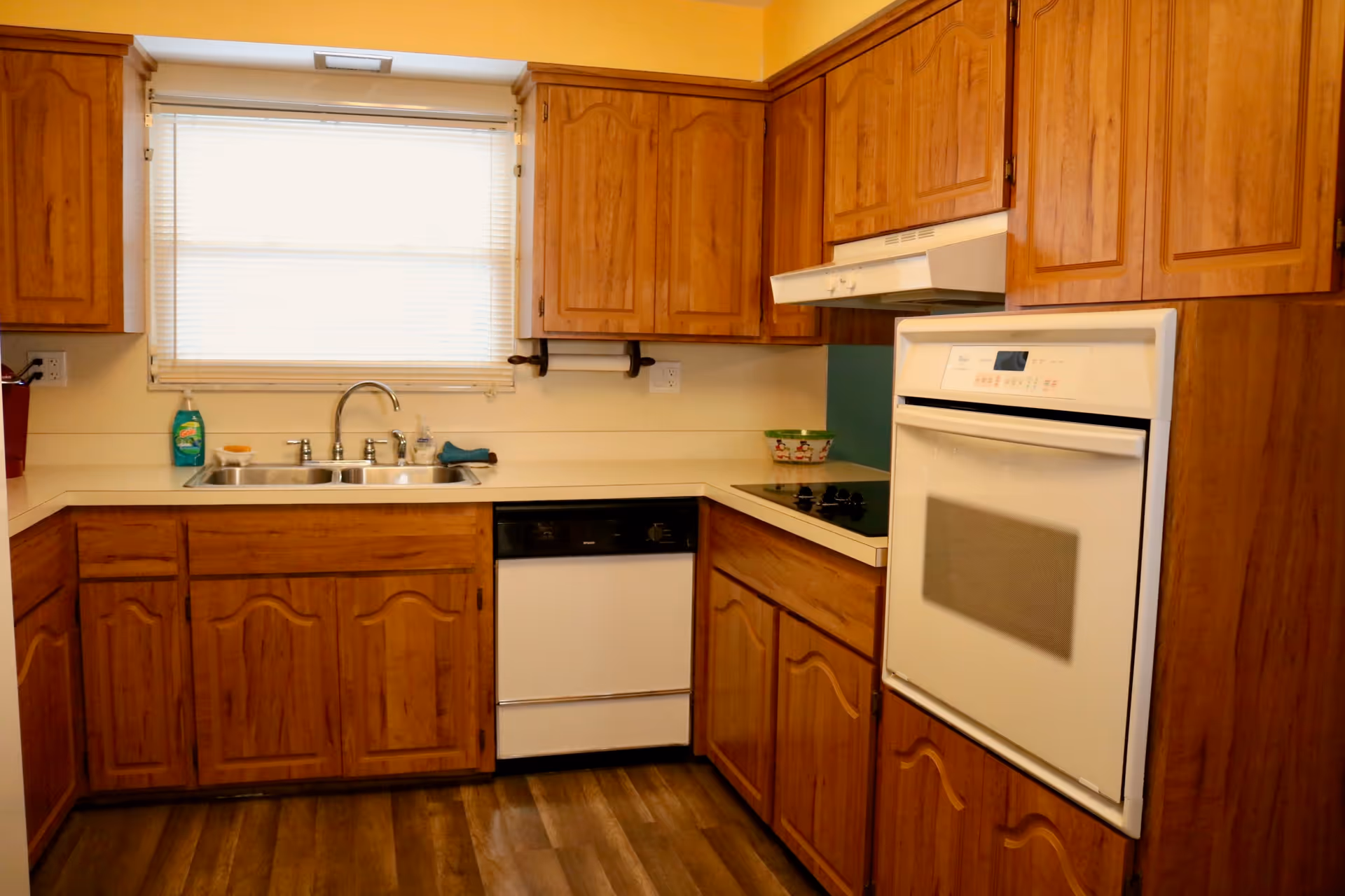 A kitchen with wooden cabinets, a double sink under a window with blinds, a dishwasher, an electric stovetop, and a built-in oven. The floor has wood-like vinyl planks.