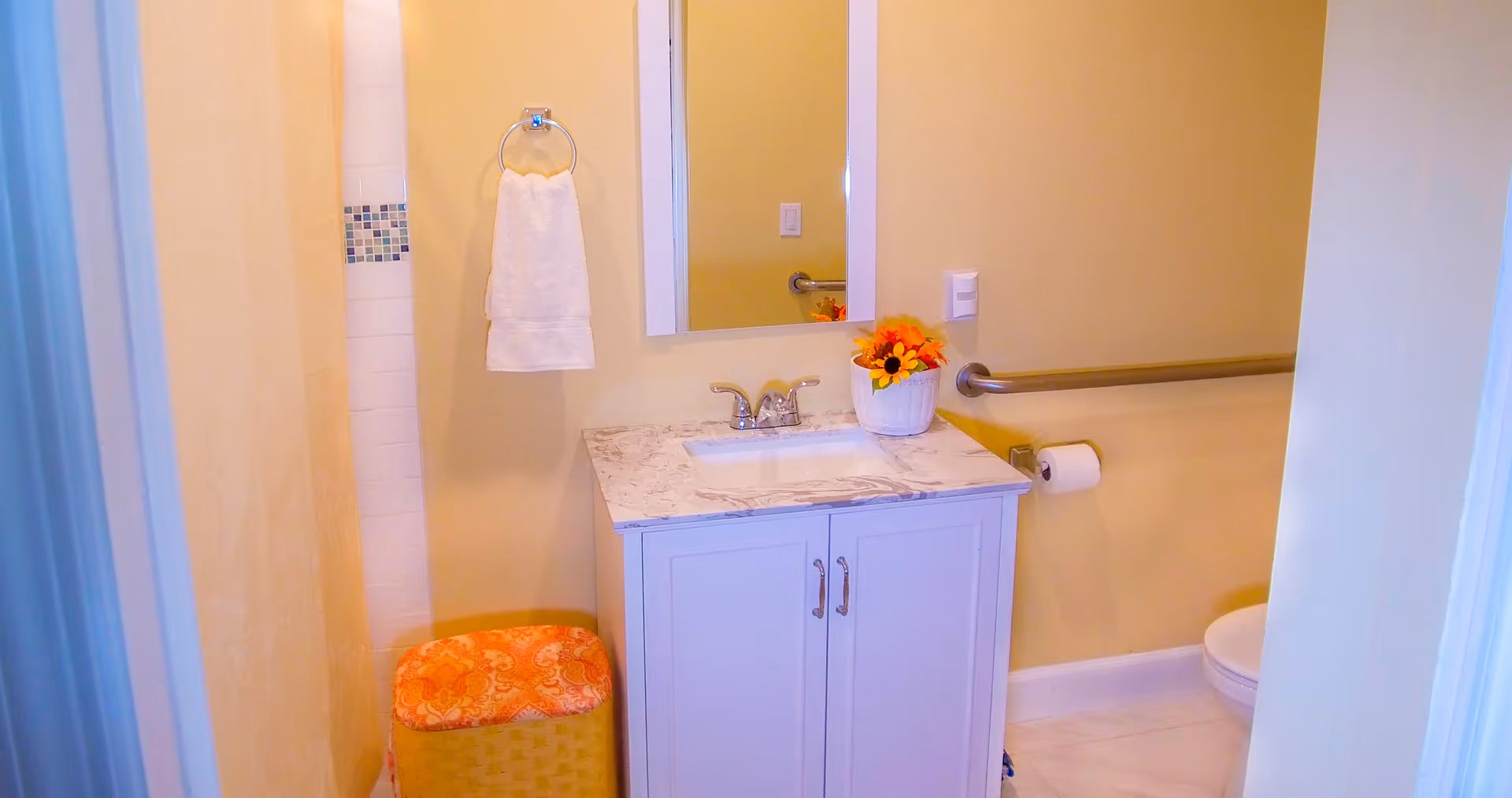 A small bathroom with a white vanity cabinet topped with a marble countertop and a sink. Above the sink is a rectangular mirror. To the left, there is a towel ring with a white towel hanging on it and a small shower area with white tiles and a decorative tile border. On the right side, there is a toilet with a grab bar mounted on the wall and a toilet paper holder. A small basket with an orange patterned cushion is placed on the floor next to the vanity. The walls are painted a light yellow color.