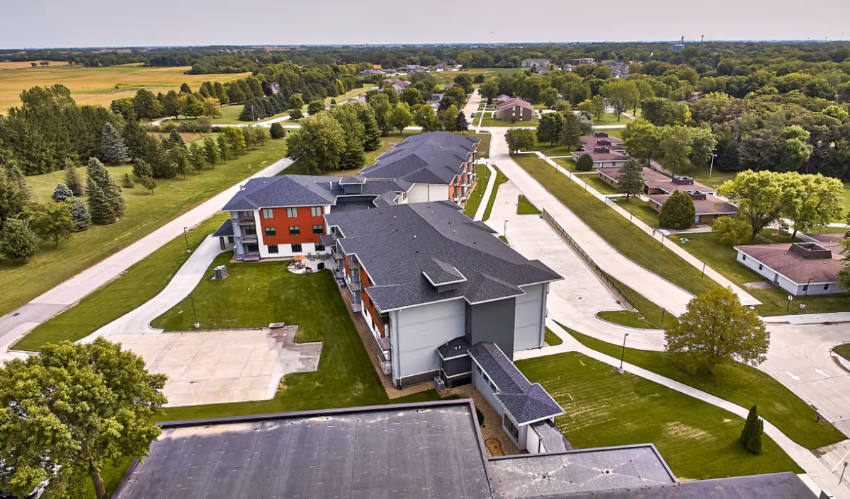 Aerial view of TimberCrest at Glen Oaks senior living facility showing multiple buildings with dark roofs surrounded by green lawns, trees, and paved roads under a clear sky.