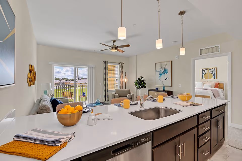 Modern open-concept living space featuring a kitchen island with a sink, dark wood cabinets, and countertop decorated with a bowl of lemons, mugs, and folded towels. In the background, there is a living room with a sofa, armchair, ceiling fan, and sliding glass doors leading to a balcony with outdoor seating. A bedroom with a bed and artwork is visible through an open doorway.