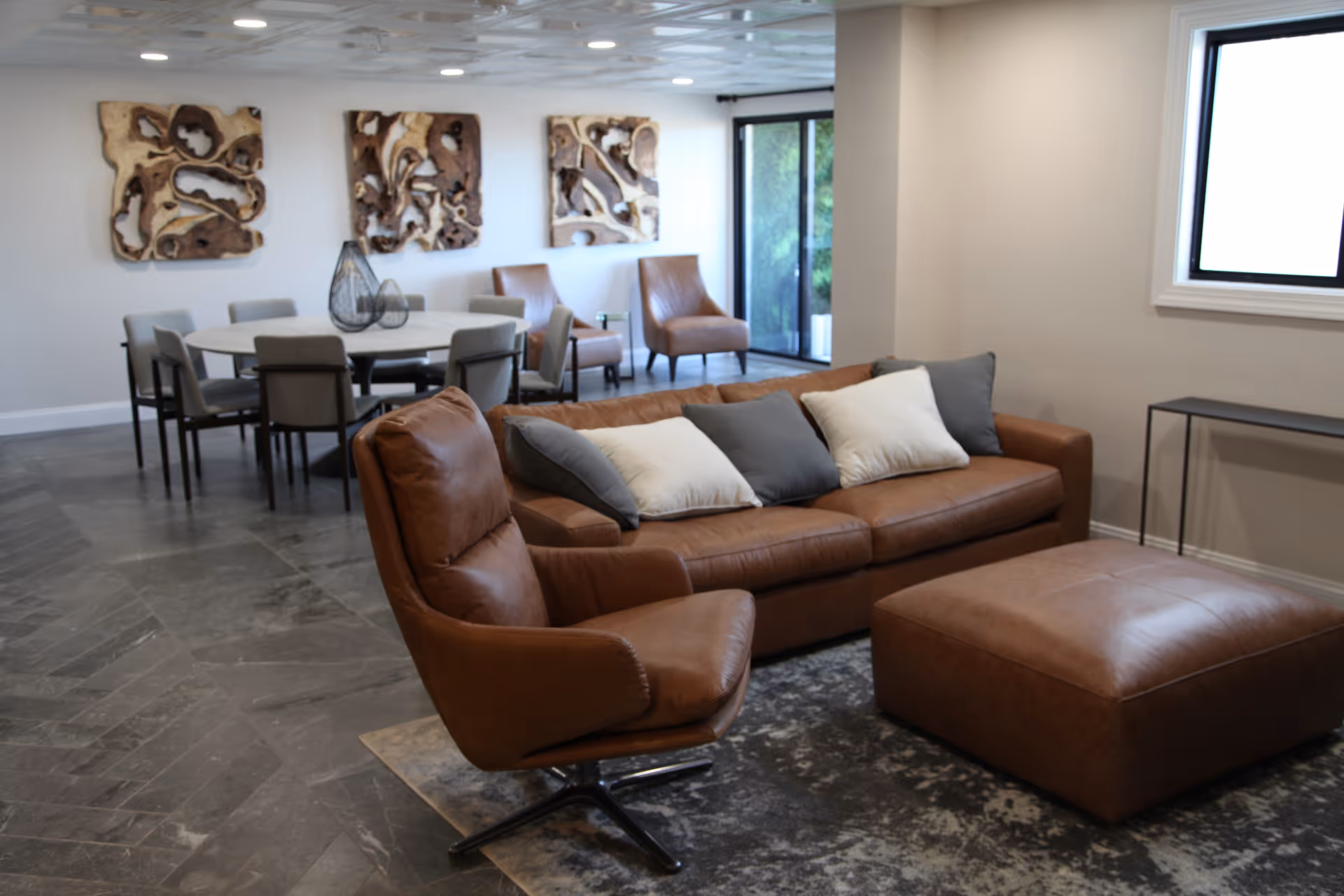 A modern living room area with a brown leather sofa adorned with white and gray pillows, a matching leather armchair, and a large leather ottoman. Behind the seating area is a dining table with several chairs and three abstract wooden wall art pieces. The room has a large window and a glass door letting in natural light.