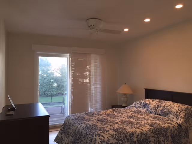 A bedroom with a bed covered in a blue and white patterned bedspread, a bedside table with a lamp, a ceiling fan, recessed lighting, a dresser with a remote control on top, and a sliding glass door with vertical blinds leading to an outdoor area with greenery.