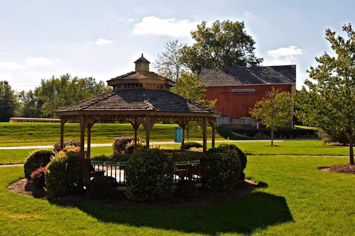 A wooden gazebo surrounded by neatly trimmed bushes and green grass in an outdoor garden area. In the background, there is a large red barn-style building and several trees under a partly cloudy sky.