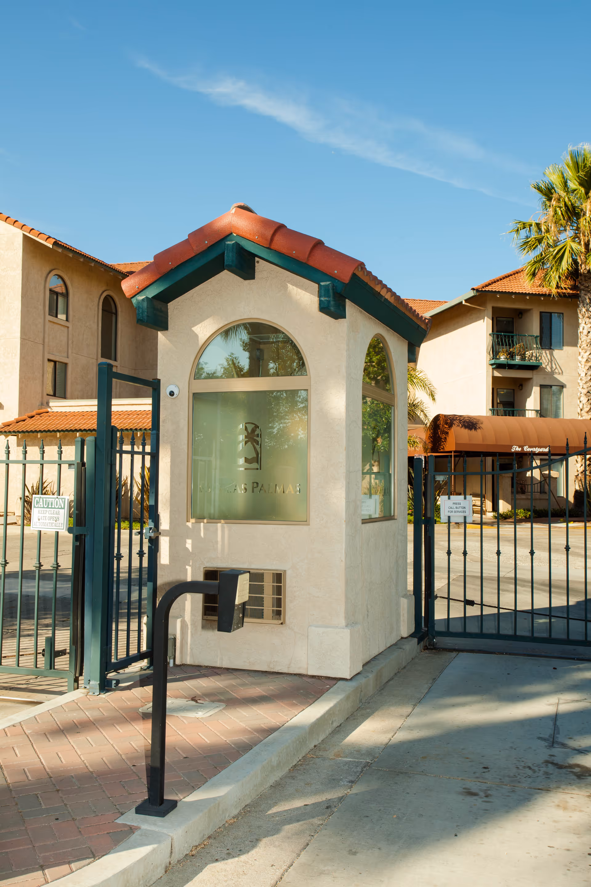 Entrance gate of Rio Las Palmas facility featuring a small guardhouse with a tiled roof and frosted windows displaying the facility's logo. The gate is black metal with a keypad entry system, and there are beige buildings and palm trees visible in the background under a clear blue sky.