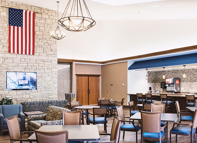 A spacious senior living facility common area with multiple tables and chairs arranged for dining or socializing. There is a stone wall with an American flag hanging and a mounted TV. Comfortable armchairs and sofas are placed near the wall. A counter with bar stools and a beverage dispenser is visible in the background under a blue awning.