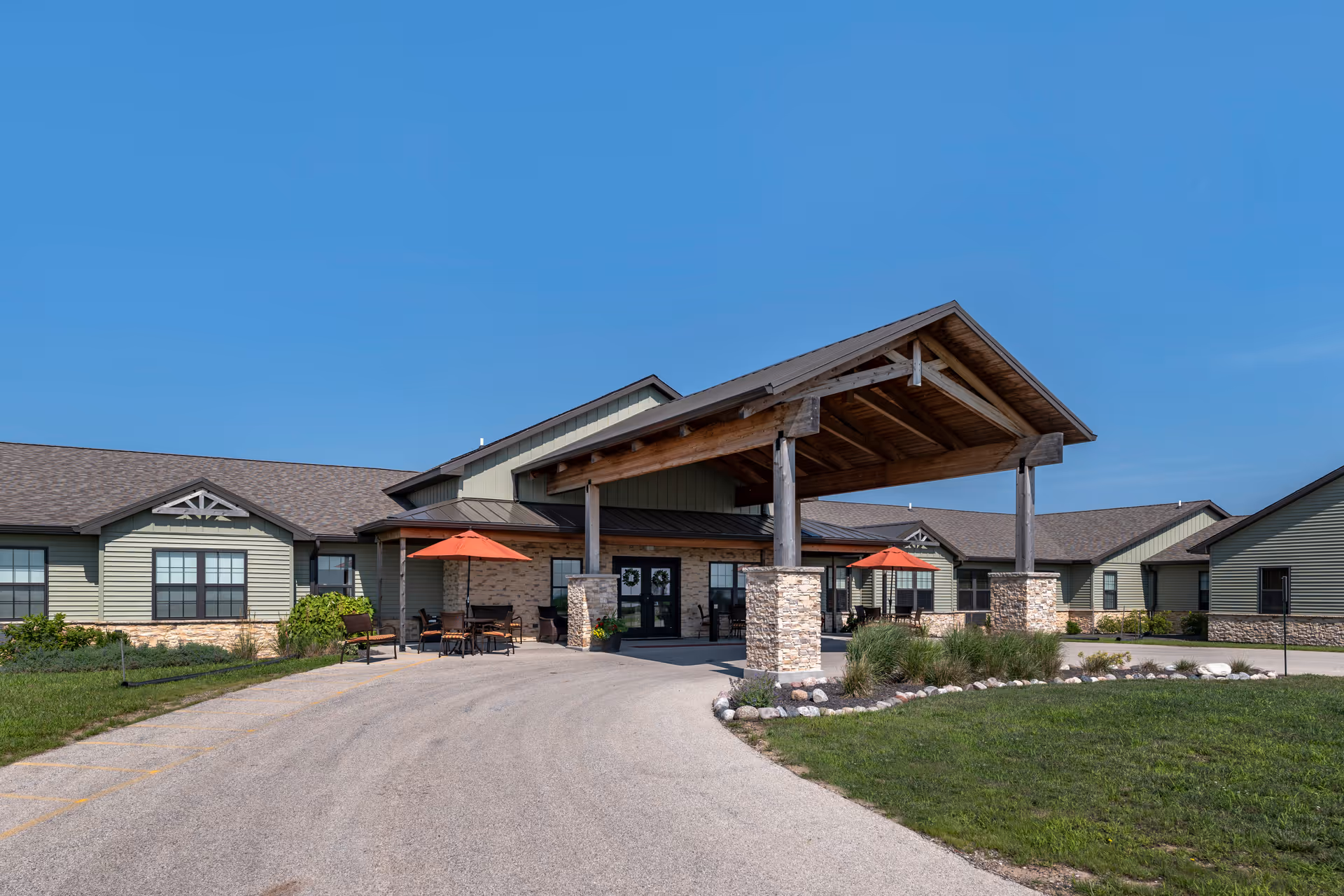 Exterior view of The Lodge at Manito senior living facility showing a single-story building with a covered entrance supported by stone pillars. There are outdoor seating areas with tables and orange umbrellas on either side of the entrance. The sky is clear and blue.