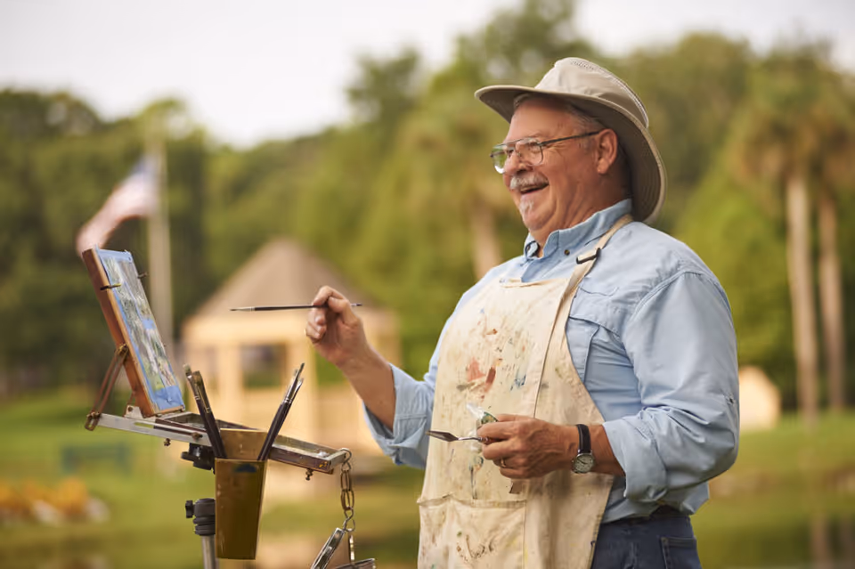 An elderly man wearing a hat and glasses, smiling while painting outdoors on an easel. He is dressed in a light blue shirt and a paint-splattered apron, with greenery and a small structure in the background.