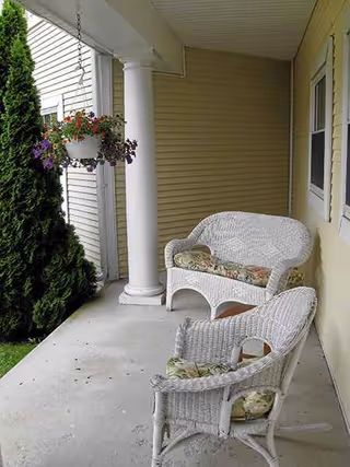 Covered outdoor porch with wicker chairs, hanging flowers, and a column beside beige siding.