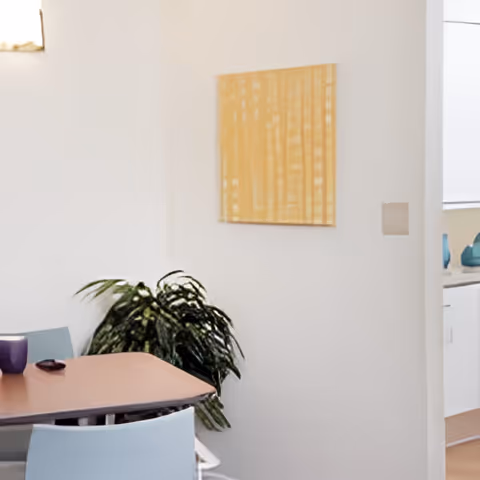 A corner of a room with a wooden table and light blue chairs, a green leafy plant in the corner, a yellow abstract painting on a white wall, and part of a kitchen area with white cabinets and blue decorative items visible.