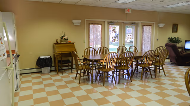 Communal dining room with a long wooden table and chairs on a checkered floor, double glass doors with curtains, and a small sitting area with a recliner and TV.