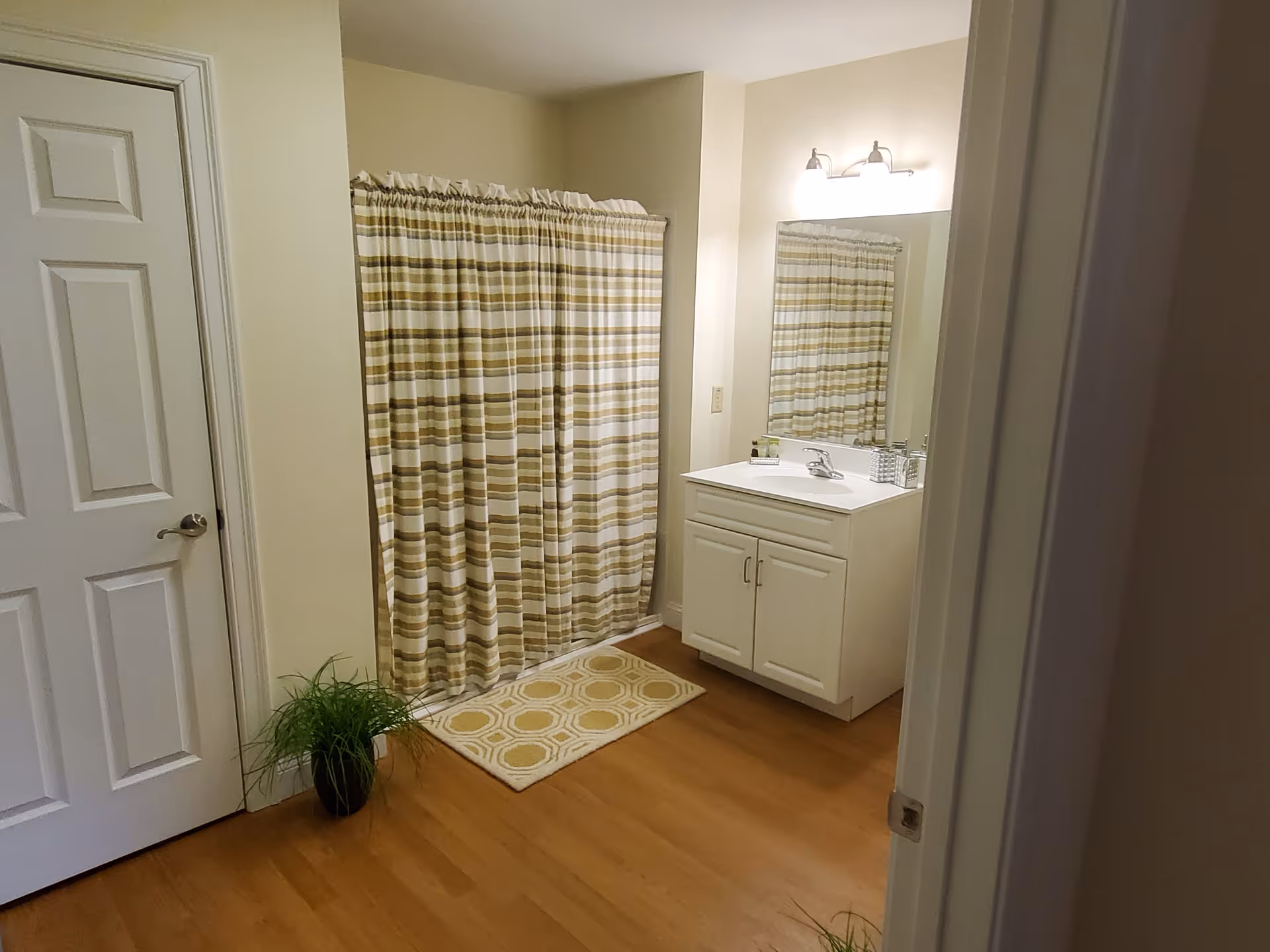A bathroom with a white door on the left, a shower with a striped beige and white curtain, a white vanity with a sink and mirror above it, a small plant on the floor, and a beige patterned bath mat in front of the shower.
