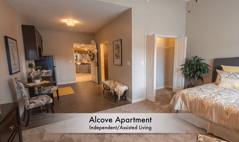 Interior view of an alcove apartment in an assisted living facility showing a bedroom area with a bed, pillows, and a plant, a small dining table with two chairs, an open closet with double doors, and a hallway leading to another room decorated with balloons.