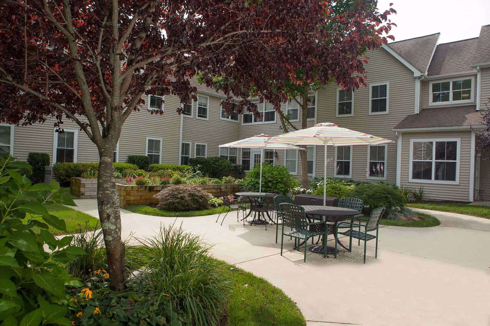 Outdoor courtyard area of a senior living facility with patio tables and chairs under striped umbrellas, surrounded by landscaped greenery and trees, with the beige exterior of the building in the background.