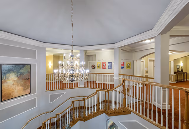 Interior view of a senior living facility featuring a wooden staircase with white railings, a large ornate chandelier hanging from the ceiling, and a hallway with framed artwork and award plaques on the walls. The area is well-lit with warm lighting and has beige and gray walls with white trim.