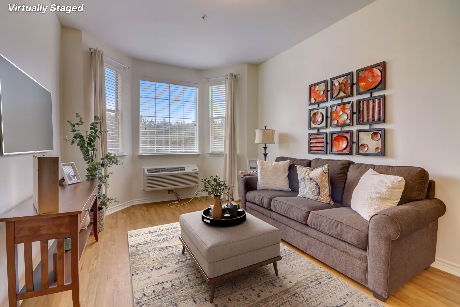 A cozy living room with a brown sofa adorned with three decorative pillows, a beige ottoman with a tray holding a vase and small decor items, a wooden console table with a framed photo and a potted plant, a wall-mounted flat-screen TV, large windows with blinds and beige curtains, and a piece of abstract wall art above the sofa.