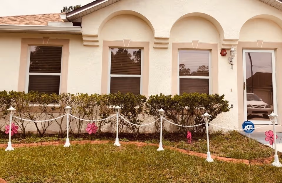 Exterior view of a light-colored building with three windows and a glass door. In front of the building, there is a small garden area with trimmed bushes, a grass lawn, and a decorative white chain fence supported by small white posts with lantern-style tops. Pink flower decorations are attached to the fence posts. A blue ADT security sign is visible near the door.