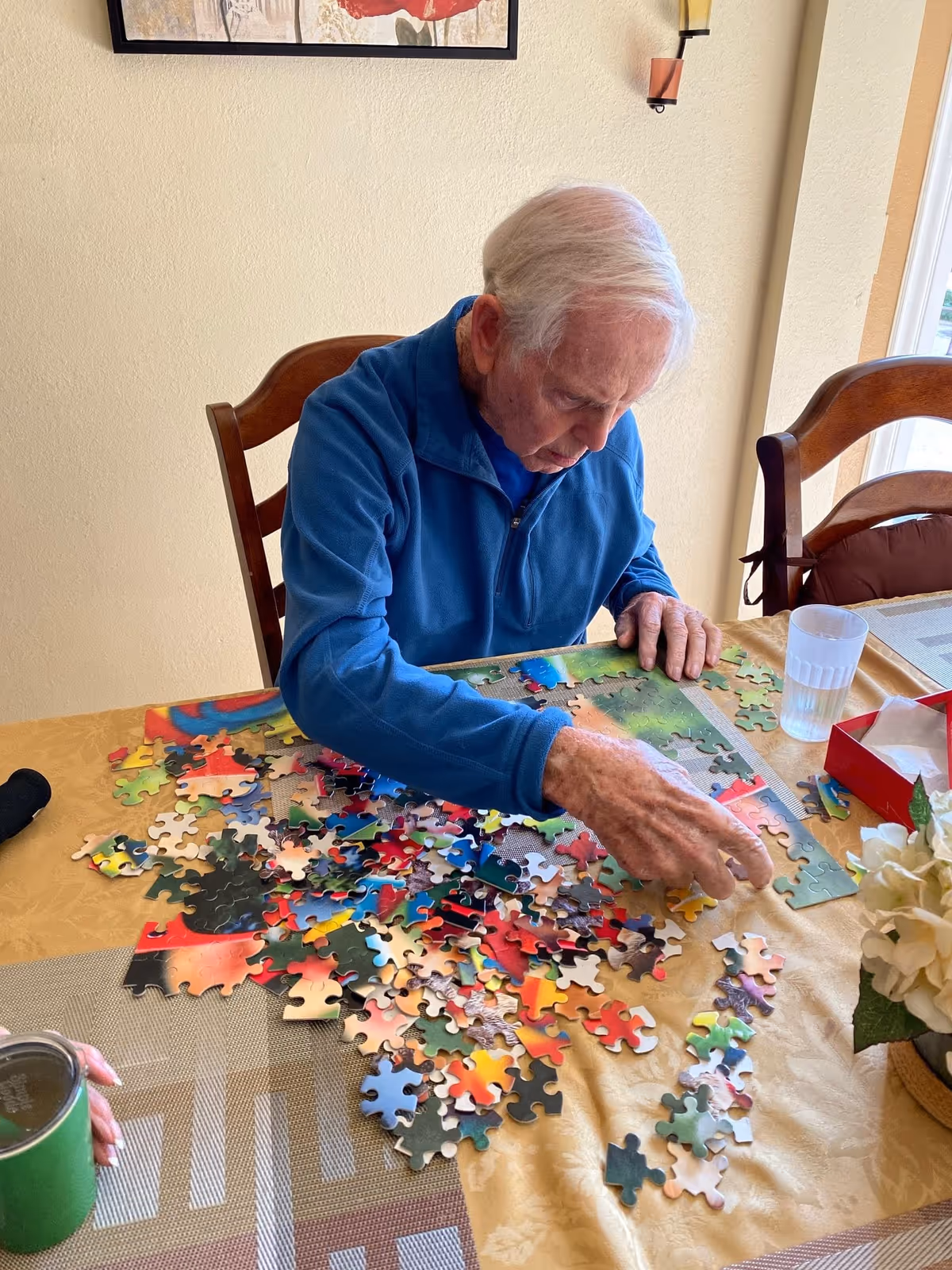 An elderly man with white hair wearing a blue jacket is sitting at a table working on assembling a colorful jigsaw puzzle. The table is covered with a yellow tablecloth and scattered puzzle pieces. There is a glass of water, a red box, and a flower arrangement on the table. The setting appears to be a well-lit room with beige walls and wooden chairs.