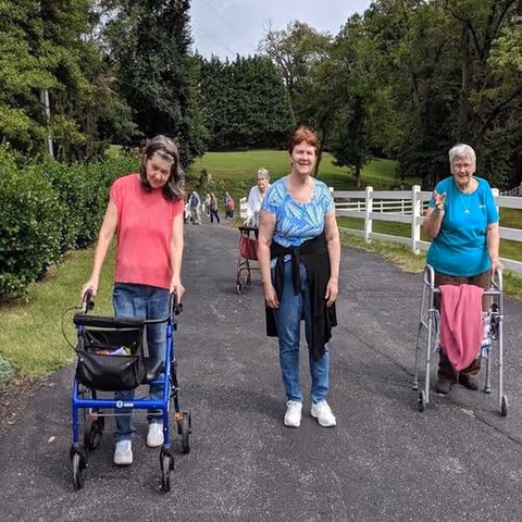 Three elderly women walking outdoors on a paved path surrounded by greenery. Two of the women are using walkers, and one woman is walking without assistance. Trees and a white fence line the path.