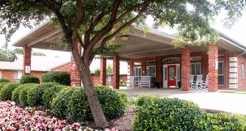 Entrance area of a senior living facility with a covered driveway supported by brick columns, surrounded by green bushes, a tree, and flower beds. The building has a red door and several white rocking chairs on the porch.