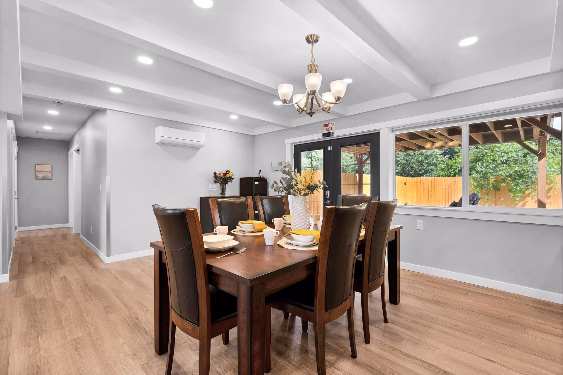 A modern dining room with a wooden table set for six people, featuring white plates, bowls, cups, and yellow napkins. The room has light wood flooring, white walls, recessed ceiling lights, and a chandelier above the table. Large windows and a glass door provide a view of a wooden fence and greenery outside.