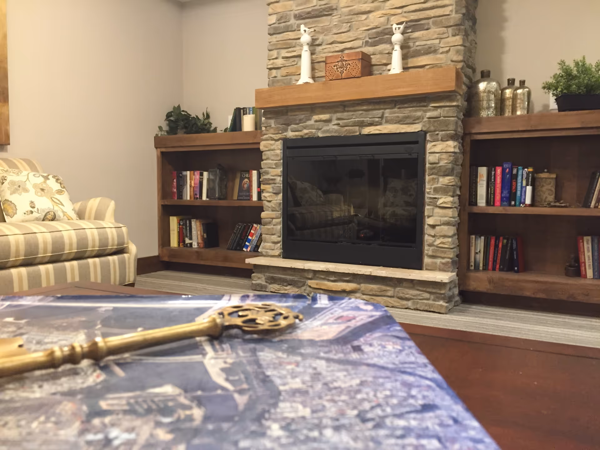 Cozy living room area with a stone fireplace in the center, flanked by wooden bookshelves filled with books and decorative items. A striped armchair with floral cushions is visible on the left side, and a table with a decorative key is in the foreground.