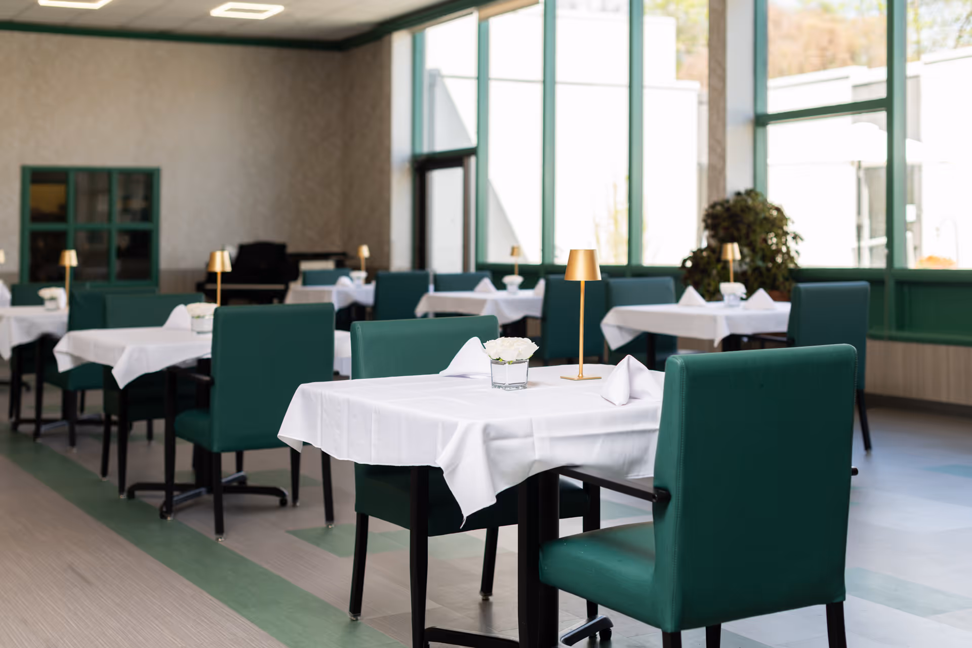 A bright dining room with multiple tables covered in white tablecloths, each set with folded napkins, small flower arrangements, and gold-colored table lamps. The room has large windows allowing natural light to fill the space, green cushioned chairs, and a plant in the corner near the windows.