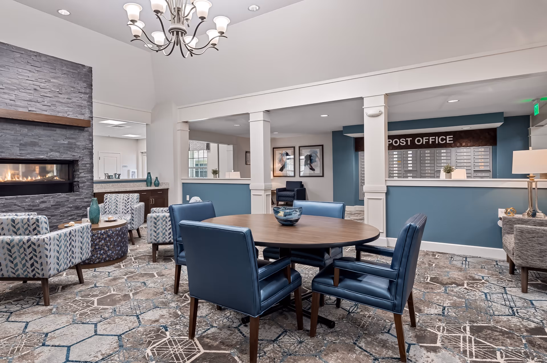 A cozy common area in a senior living facility featuring a round wooden table with four blue leather chairs, patterned armchairs near a modern stone fireplace, and a post office section with mailboxes in the background. The room has a patterned carpet, blue and white walls, and a chandelier hanging from the ceiling.