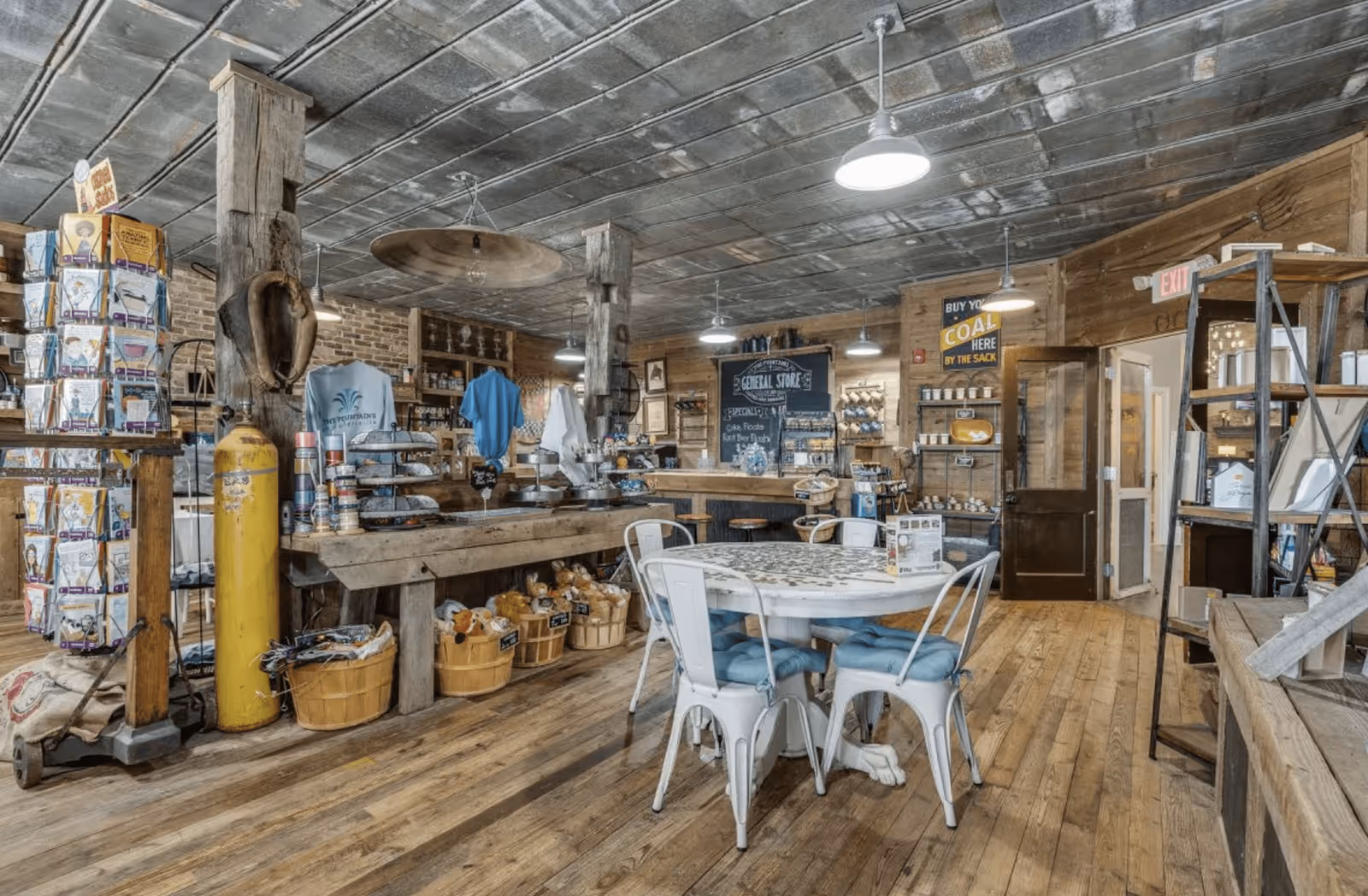 Interior of a rustic general store with wooden floors, exposed brick walls, and a tin ceiling. There are wooden shelves and tables displaying various goods including baskets, canned items, and clothing. A round table with four metal chairs with blue cushions is in the foreground. Hanging lights illuminate the space, and signs advertise products like coal.