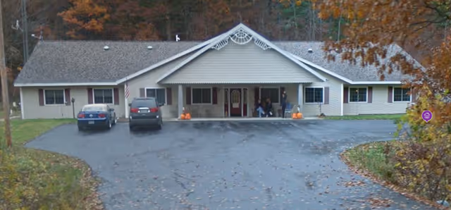Front exterior view of a single-story building with a gabled roof, beige siding, and a covered entrance. Two cars are parked in front on a paved driveway. There are a few people sitting near the entrance, and autumn trees with orange leaves surround the building.