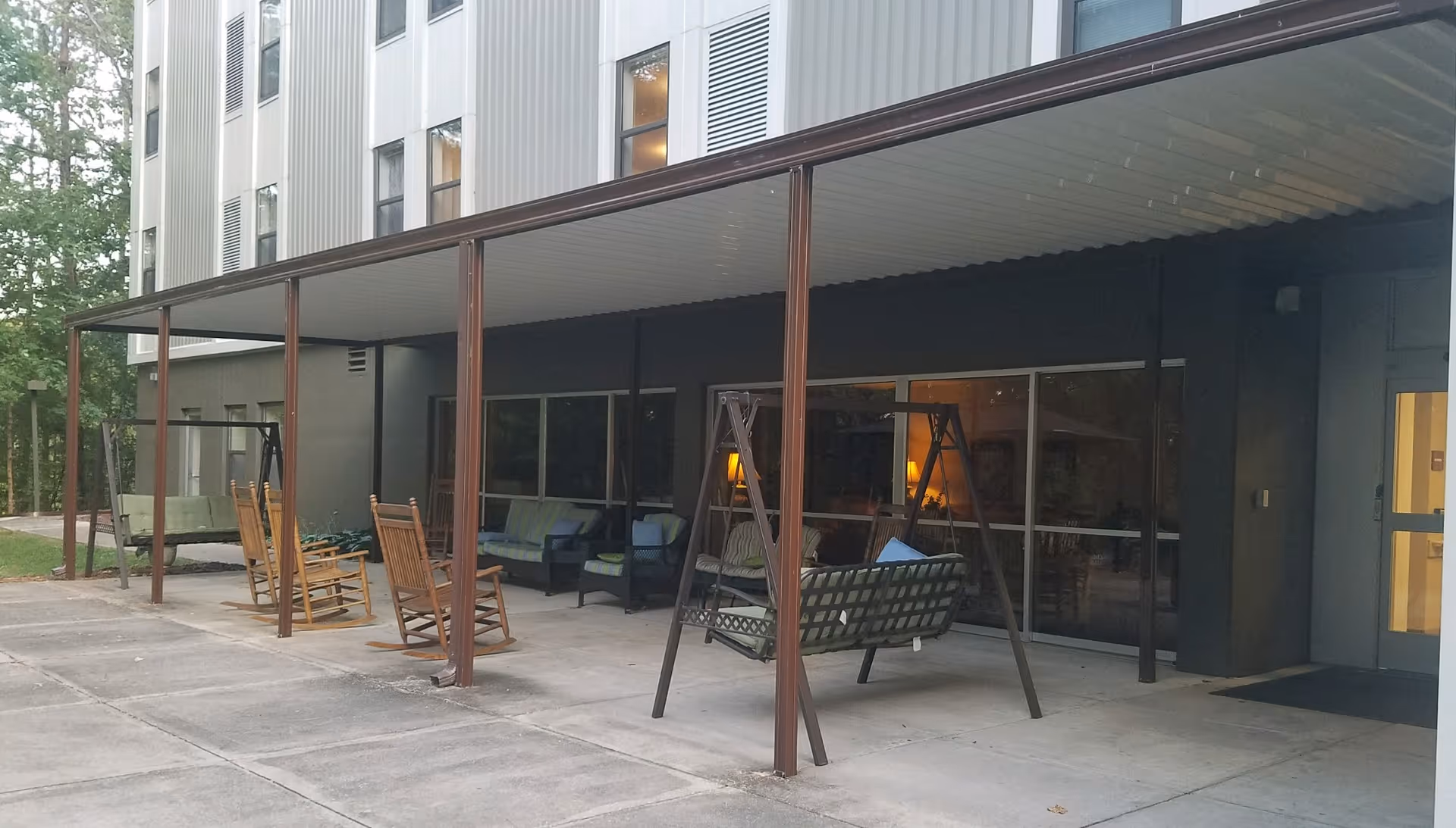 Covered outdoor patio area with porch swings, rocking chairs and cushioned seating along a building façade.