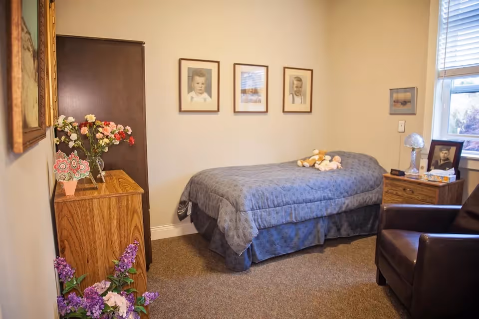 A small bedroom with a single bed covered in a blue patterned bedspread. On the bed are two stuffed animals. Above the bed, three framed pictures hang on the wall. To the right of the bed is a wooden nightstand with a lamp, a framed photo, and a box of tissues. A window with blinds is above the nightstand. On the left side of the room, there is a wooden dresser with a vase of flowers and decorative items on top. A dark wooden wardrobe stands next to the dresser. The floor is carpeted, and there is a brown leather armchair in the foreground.