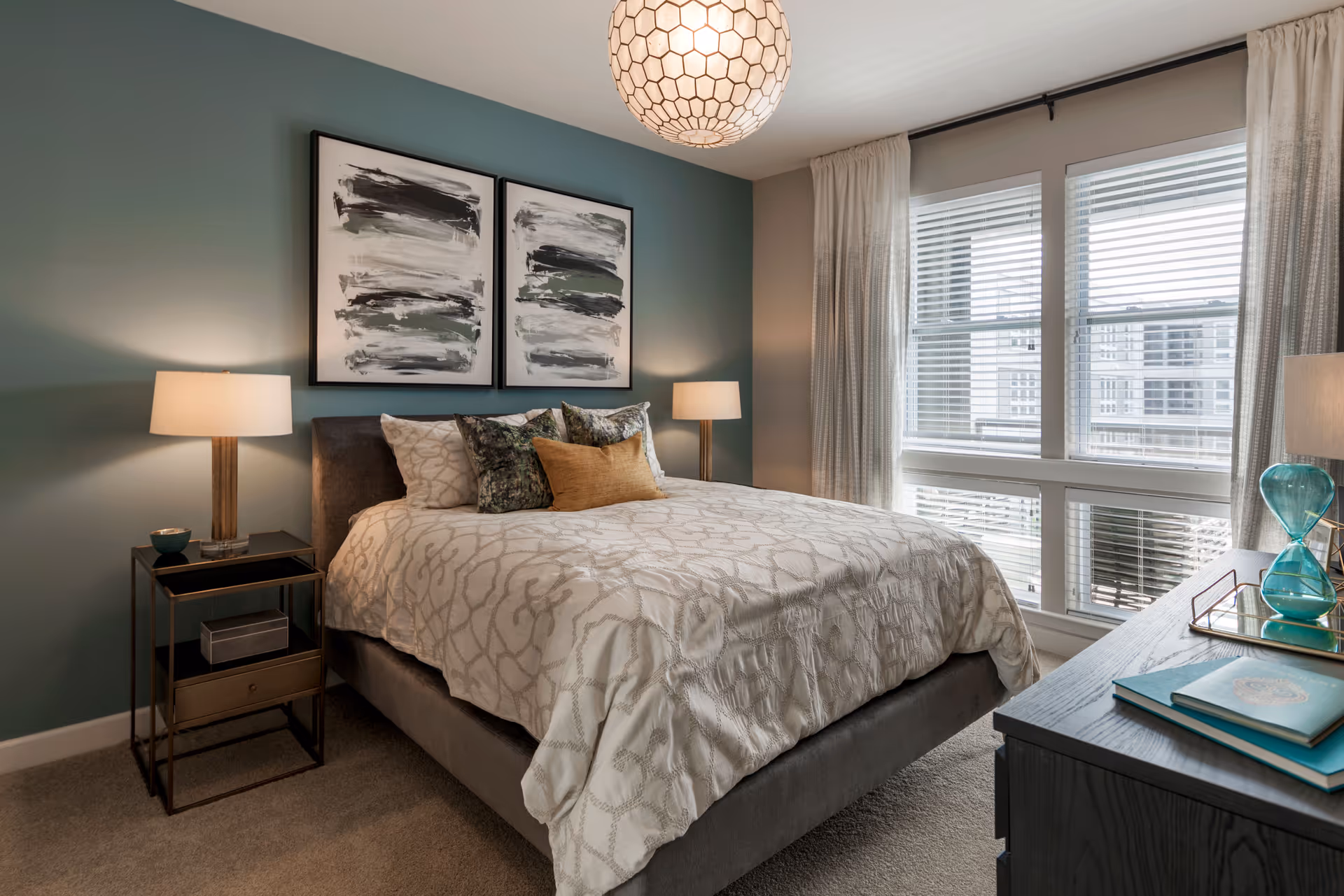 Bedroom with a made bed, two bedside lamps, abstract artwork above the headboard, and a large window with blinds.
