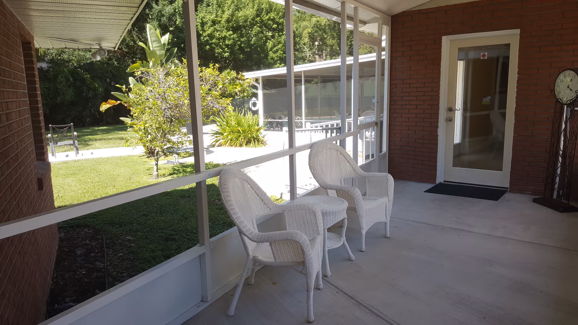 A screened-in porch area with two white wicker chairs and a small matching table between them. The porch overlooks a green lawn with trees and bushes. There is a brick wall with a glass door and a clock mounted on a stand to the right.