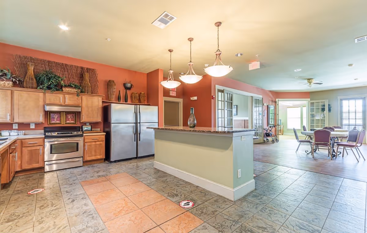 A spacious kitchen area with wooden cabinets, stainless steel refrigerator and oven, and a countertop island with a decorative vase. The kitchen opens into a dining area with tables and chairs, and large windows letting in natural light.