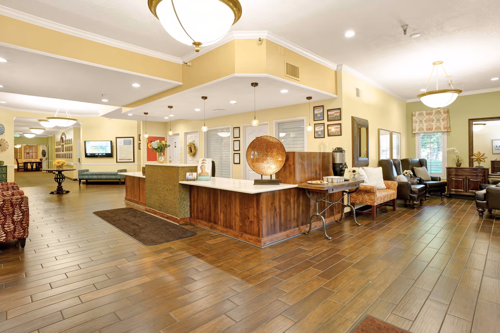 A spacious and well-lit reception area in a senior living facility with a wooden front desk, pendant lights, comfortable seating including armchairs and sofas, decorative wall art, and a coffee station with cups and cookies on a side table.